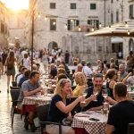 Crowded old-town square with tourists at outdoor cafes under string lights on a summer evening