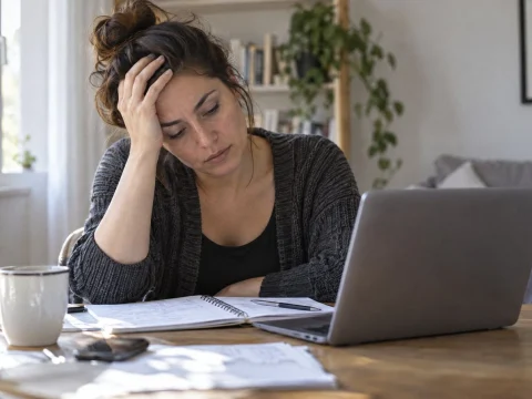 Tired woman at a home office desk holding her head beside a laptop and notes