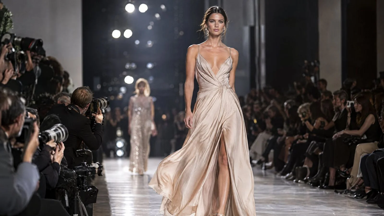 Model walking a runway in an elegant outfit while photographers line the catwalk under bright show lights