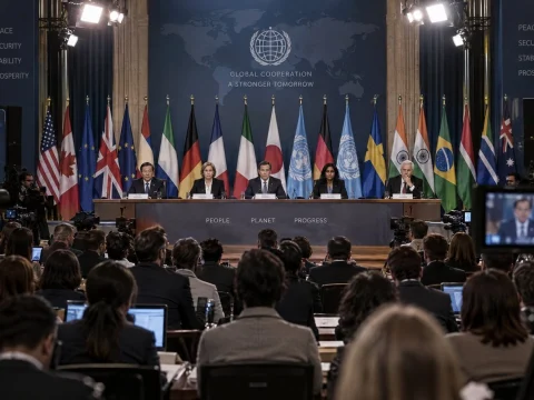 Leaders seated behind a long table in front of many national flags at a press conference, cameras in the foreground