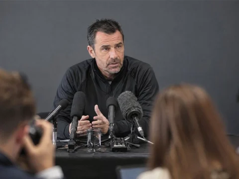 Football coach speaking into microphones at a press conference table, journalists in the foreground
