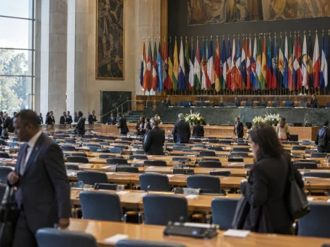 Wide shot of an assembly hall with rows of empty delegate seats and flags at the back