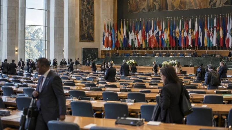 Wide shot of an assembly hall with rows of empty delegate seats and flags at the back