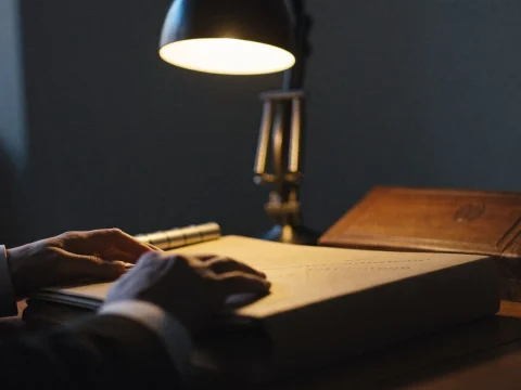 An official's hands resting on a thick closed folder on a wooden desk under a warm desk lamp