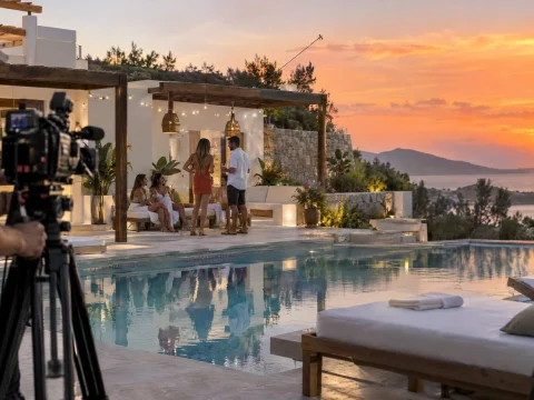 Reality TV villa with a pool and sun loungers at sunset, cameras on tripods at the edge of the terrace