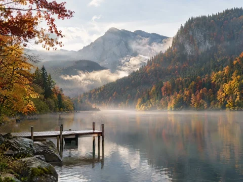 Mountain lake surrounded by red and gold autumn forest, with a wooden pier in the morning mist