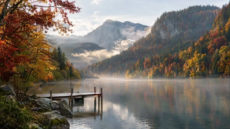 Mountain lake surrounded by red and gold autumn forest, with a wooden pier in the morning mist