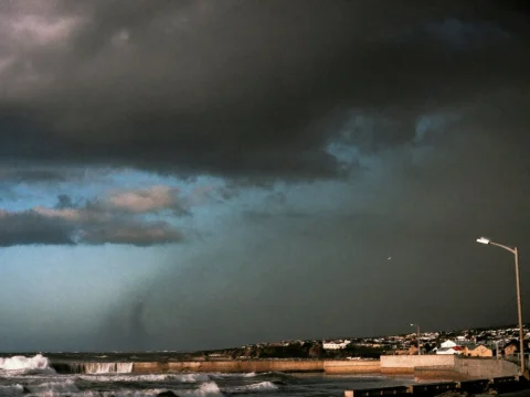 Dark storm clouds over a coastal town as waves crash against a seawall in heavy rain