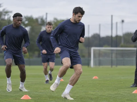 Football players in plain training kits warming up among cones on a training pitch while a coach holds a whistle