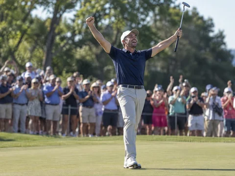 Golfer raising both arms to celebrate a putt on the green, with spectators behind the ropes on a sunny day
