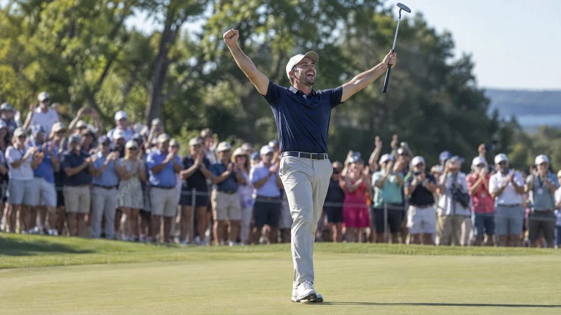 Golfer raising both arms to celebrate a putt on the green, with spectators behind the ropes on a sunny day