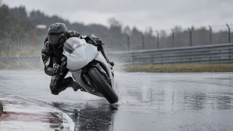 Racing motorcycle leaning into a corner on a wet track, throwing up spray under a grey sky