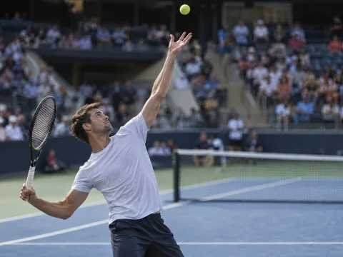 Tennis player frozen mid-serve on a hard court inside a stadium, wearing plain clothing