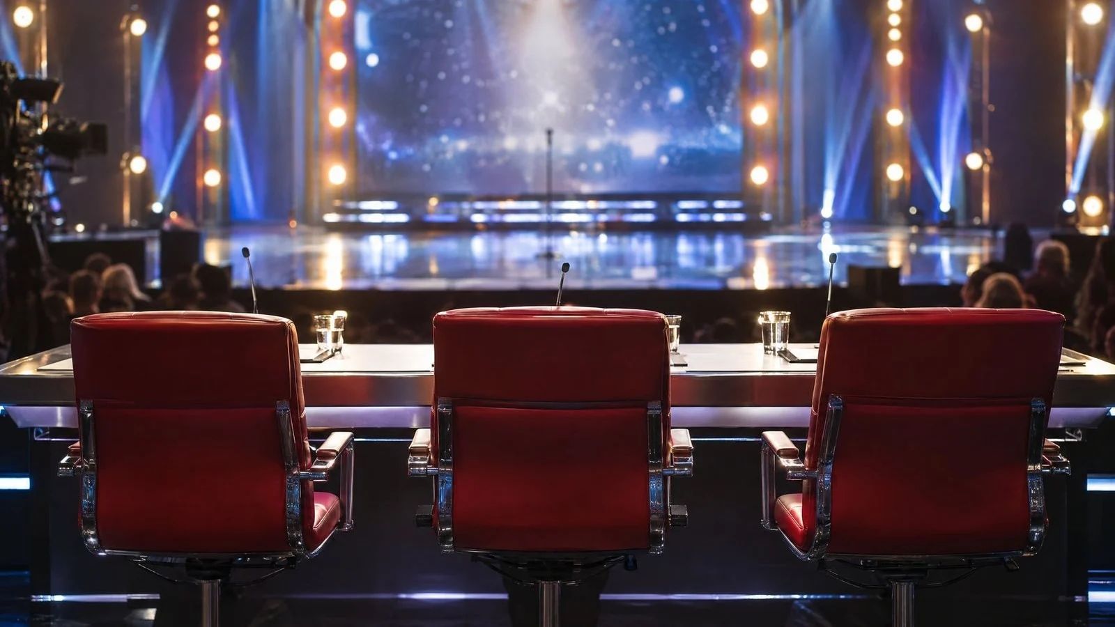 Talent show judging desk with three empty red chairs facing a lit stage in a TV studio