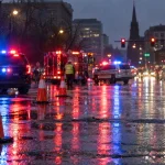 Rainy city intersection at night with blurred blue and red emergency lights on wet asphalt and traffic cones