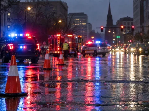 Rainy city intersection at night with blurred blue and red emergency lights on wet asphalt and traffic cones