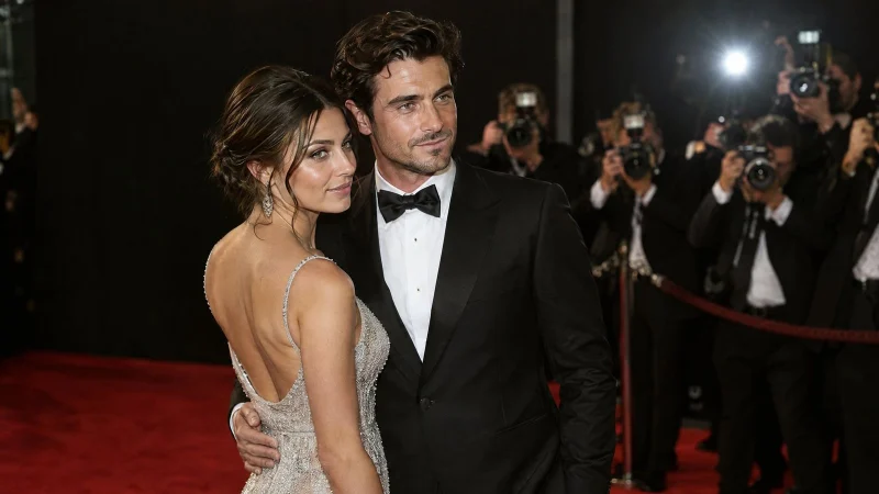 Photographers with flashing cameras in front of a glamorous couple on a red carpet against a dark backdrop